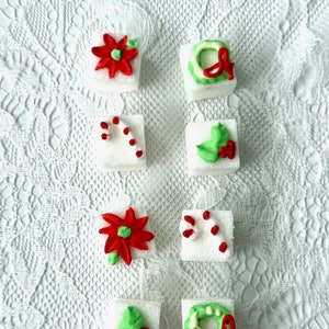 May include: Eight white sugar cubes decorated with red and green icing. The designs include a poinsettia, a candy cane, a wreath, and holly.
