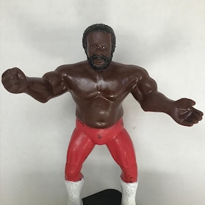 May include: A vintage action figure of a wrestler with a black afro, brown skin, and a red wrestling outfit. The figure is posed with arms outstretched and is standing on a black base.