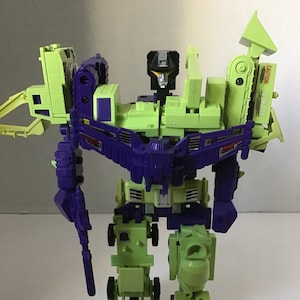 May include: A green and purple robot toy in robot mode. The robot has a construction vehicle theme and has a yellow and black head. The robot has a large, purple weapon in its right hand. The robot is standing on a white surface.