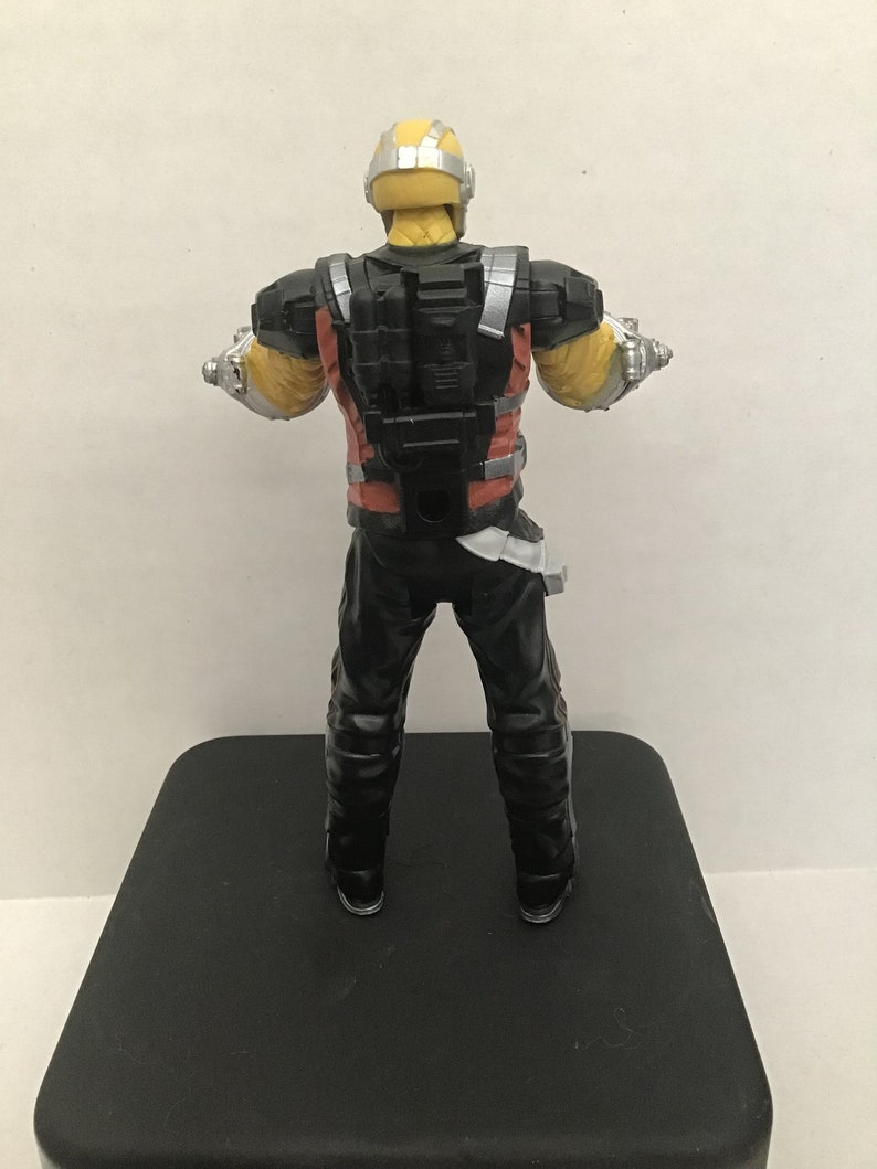 May include: A black and red action figure with a yellow helmet and silver accents. The figure is wearing a black and red suit with a black backpack. The figure is standing on a black surface.