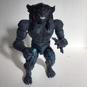 May include: A dark gray, muscular action figure with a beast-like appearance, featuring textured fur detailing on the upper body, sharp fangs, yellow eyes, and metallic blue-tinted leg armor. The figure has articulated joints and long, dark hair.