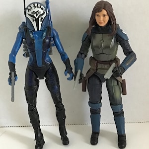 May include: Two action figures of characters in combat gear. One figure is in blue and black armor with a helmet. The other figure is in gray and black armor. Both figures are holding weapons and standing on a white surface.