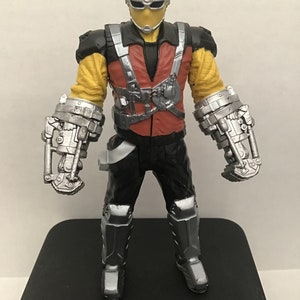 May include: Action figure of a superhero with a yellow and black suit, a red vest, and silver robotic arms. The figure has a black mask with a silver visor.