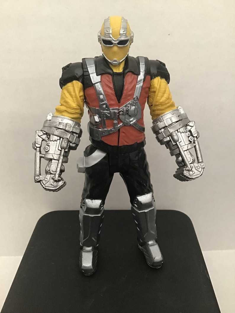 May include: Action figure of a superhero with a yellow and black suit, a red vest, and silver robotic arms. The figure has a black mask with a silver visor.