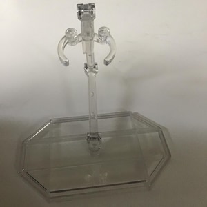 May include: A clear plastic display stand with a rectangular base and a vertical support. The stand has a series of curved arms and a top clip, designed to hold an item. The stand is made of transparent plastic.