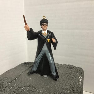 May include: A figurine of Harry Potter in his Hogwarts uniform, holding a wand. The figure wears a black robe, gray pants, and a yellow tie. The ornament has a gold loop for hanging.