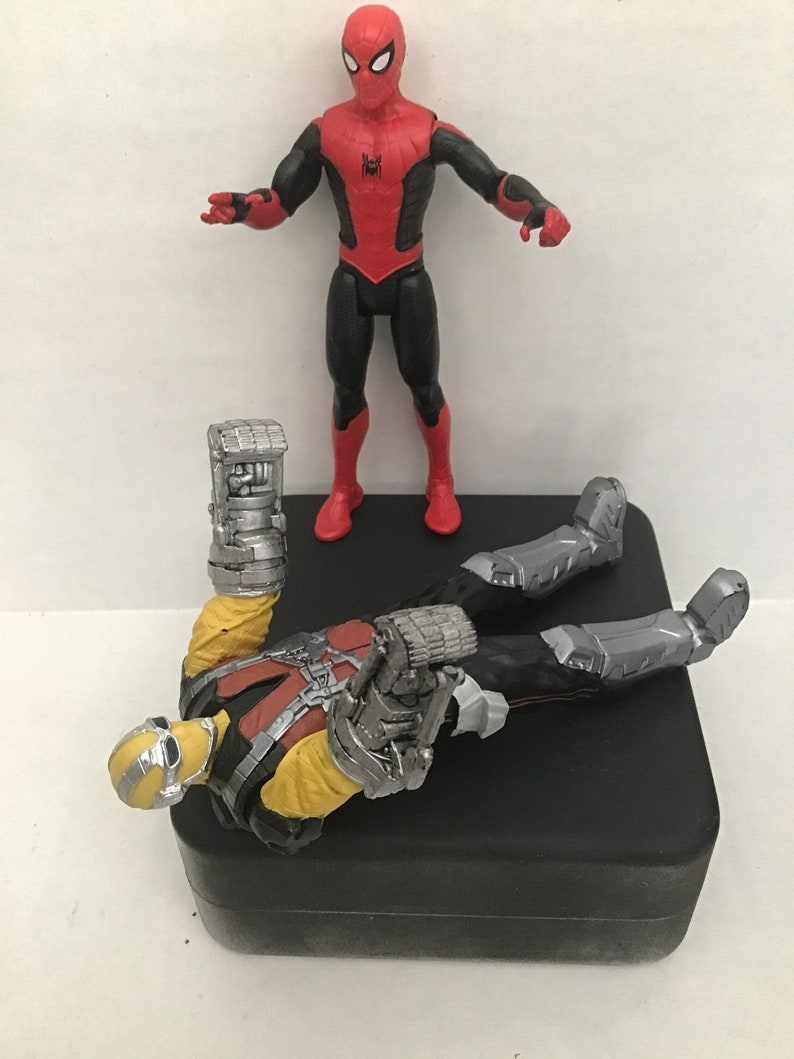 May include: A red and black Spider-Man action figure stands over a yellow and silver action figure lying on its back on a black base.