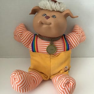 May include: A plush toy dog with orange and white striped clothing, a yellow hat, and a gold tag that says "#322".