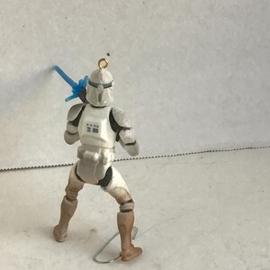 Puede incluir: Figura de acci&oacute;n blanca y gris de un soldado con un arma azul. La figura est&aacute; en posici&oacute;n de combate, con un lazo dorado en la parte superior para colgar. La figura lleva un casco y una armadura blancos.