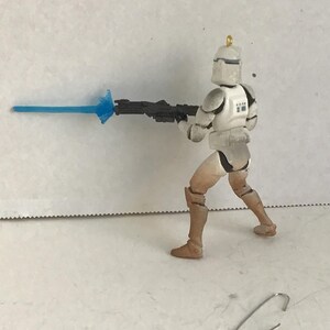 Puede incluir: Figura de acci&oacute;n de un soldado clon de Star Wars en posici&oacute;n de combate, sosteniendo un rifle bl&aacute;ster negro con una explosi&oacute;n de energ&iacute;a azul. La figura es principalmente blanca y beige, con armadura detallada y un casco. Un clip plateado est&aacute; en la superficie.