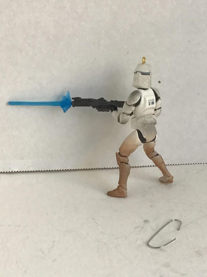 Puede incluir: Figura de acci&oacute;n de un soldado clon de Star Wars en posici&oacute;n de combate, sosteniendo un rifle bl&aacute;ster negro con una explosi&oacute;n de energ&iacute;a azul. La figura es principalmente blanca y beige, con armadura detallada y un casco. Un clip plateado est&aacute; en la superficie.