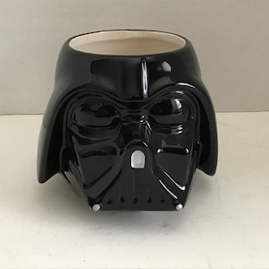 May include: A glossy black ceramic mug shaped like the helmet of Darth Vader. The mug features a smooth, reflective surface with molded details representing the iconic mask, including the triangular nose piece and vents, with a cream-colored interior.