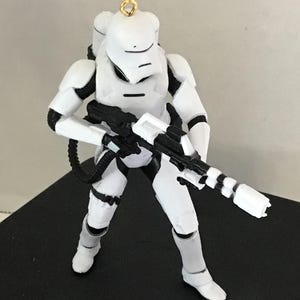 May include: White plastic Star Wars ornament of a First Order Stormtrooper with a black blaster rifle. The ornament has a gold loop for hanging.