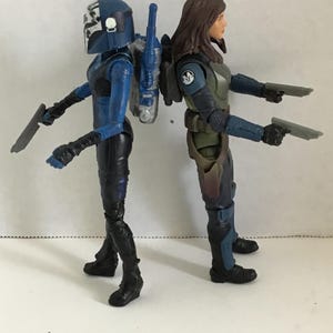 May include: Two action figures of female characters, standing back-to-back. One figure wears a blue helmet and armor, holding a silver blade. The other figure wears green and blue armor and holds two silver pistols. Both figures have black boots.