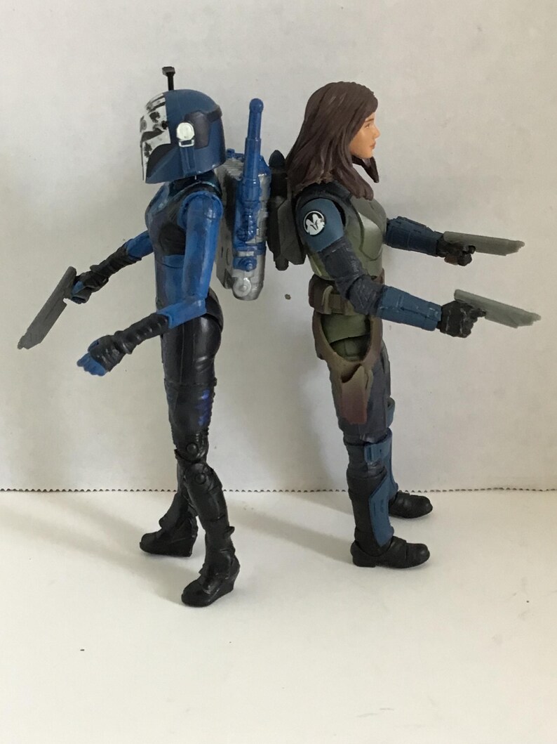 May include: Two action figures of female characters, standing back-to-back. One figure wears a blue helmet and armor, holding a silver blade. The other figure wears green and blue armor and holds two silver pistols. Both figures have black boots.
