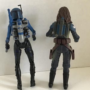 May include: Two action figures, viewed from the back, stand side-by-side. The figure on the left is blue and black, with a helmet and jetpack. The figure on the right is wearing a dark green and black outfit with a jetpack and brown belt.