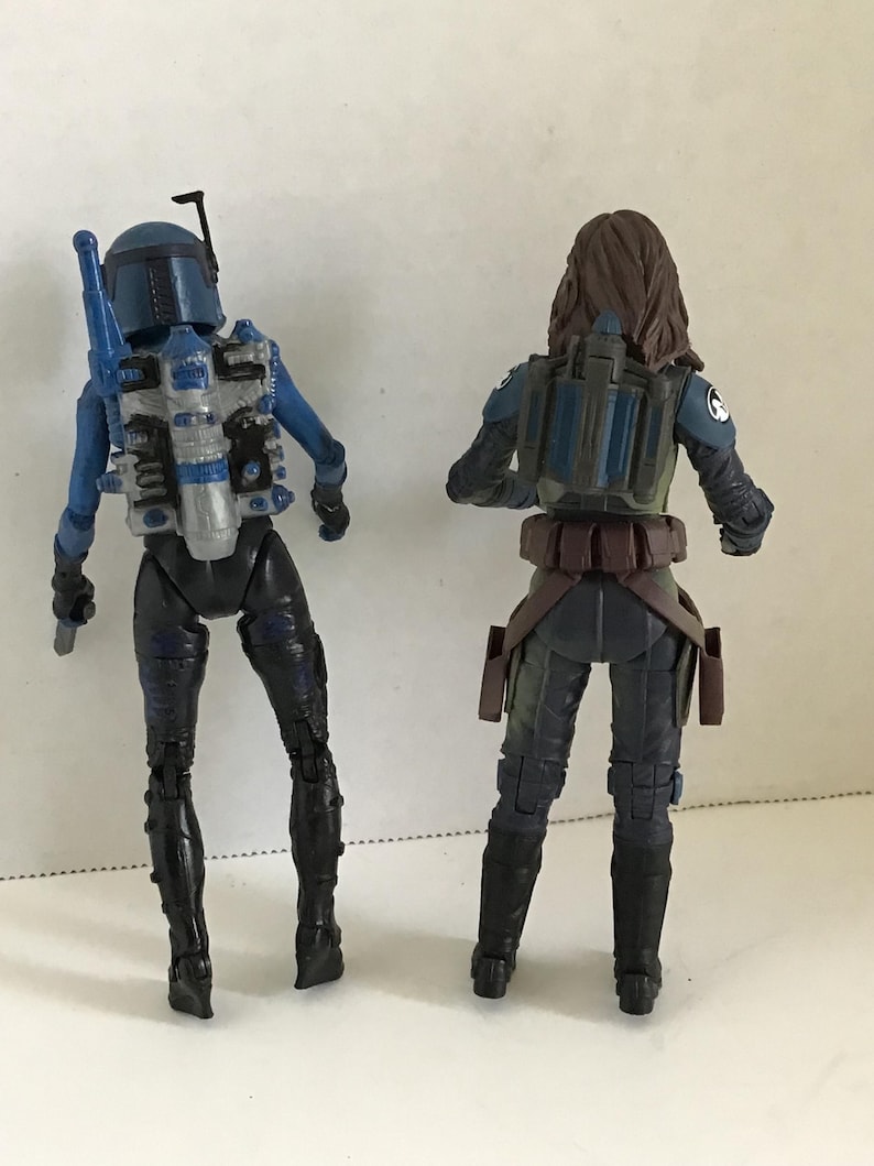 May include: Two action figures, viewed from the back, stand side-by-side. The figure on the left is blue and black, with a helmet and jetpack. The figure on the right is wearing a dark green and black outfit with a jetpack and brown belt.