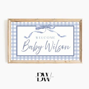 May include: A framed sign with a light wood border. The sign features a blue gingham border and a white center with the words "WELCOME Baby Wilson" in elegant script, topped with a blue ribbon.