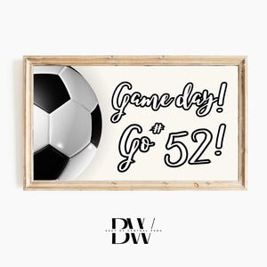 May include: A framed print featuring a soccer ball graphic and the words "Game day! Go #52!" in black script. The soccer ball is black and white. The frame is a light wood colour.