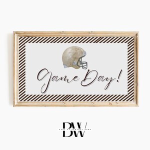 May include: A framed print with a light background, featuring a football helmet illustration and the words "Game Day!" in elegant script. The frame is a light wood color, and the print has a brown and white striped border.