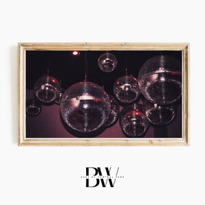 May include: A framed print featuring multiple disco balls hanging from a dark ceiling. The mirrored spheres reflect light, creating a sparkling effect. The frame is a light wood color. The image has a vintage, retro aesthetic.