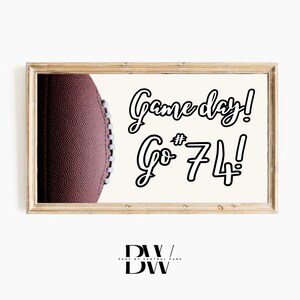 May include: A rectangular framed print featuring a textured brown football on the left side and the black text Game day! Go # 74! on a cream background. The frame is made of light-coloured wood with a visible grain, surrounding the artwork.