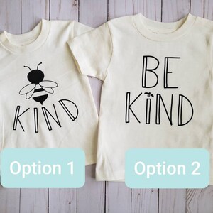May include: Two white t-shirts with black text. The first shirt has a bee and the word "KIND". The second shirt says "BE KIND".