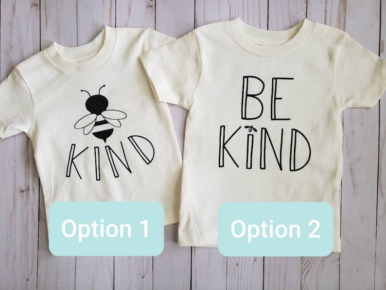 May include: Two white t-shirts with black text. The first shirt has a bee and the word "KIND". The second shirt says "BE KIND".