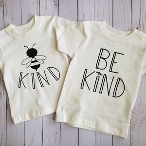 May include: Two white t-shirts with short sleeves. One shirt has a black and white bee graphic and the text "KIND". The other shirt has the text "BE KIND" in black.