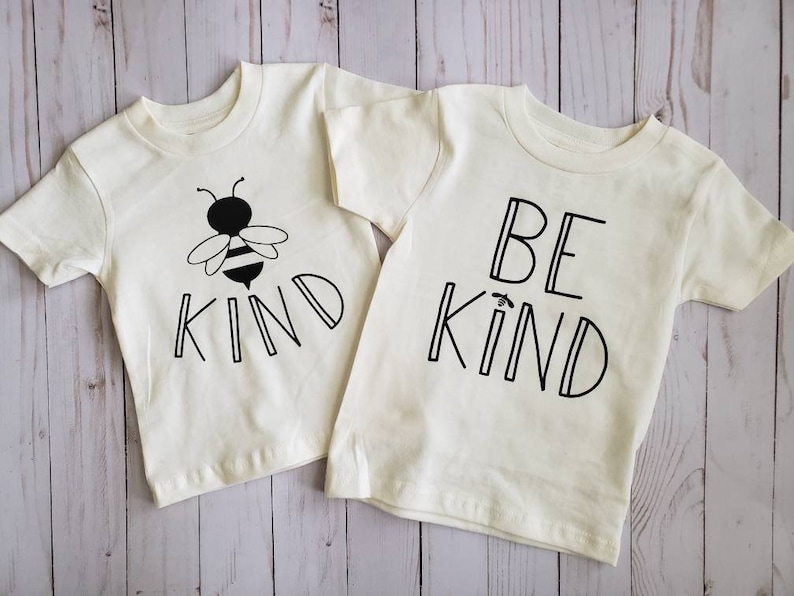 May include: Two white t-shirts with short sleeves. One shirt has a black and white bee graphic and the text "KIND". The other shirt has the text "BE KIND" in black.