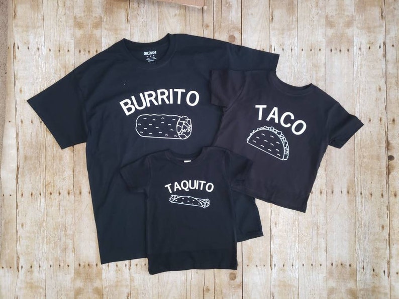 May include: Three black t-shirts with white text and illustrations. The largest shirt says "Burrito" with a burrito illustration. The medium shirt says "Taco" with a taco illustration. The smallest shirt says "Taquito" with a taquito illustration.
