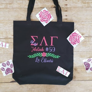 May include: Black tote bag with a white design that says "ΣΛΓ Idaliah #59 La Estuari" with a floral design and paw prints. The bag is surrounded by white stickers with the letters "ΣΛΓ" and the numbers "1990" on them.