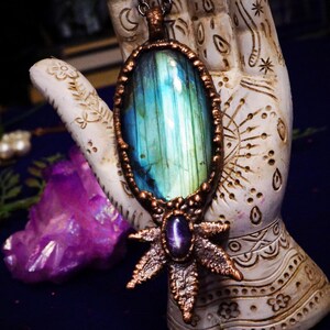 May include: A copper pendant with a large iridescent blue labradorite gemstone and a smaller amethyst stone in the center of a copper starburst design. The pendant is hanging from a silver chain.