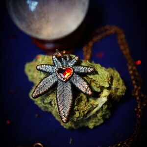 May include: A silver-toned pendant necklace featuring a stylized cannabis leaf with a red heart-shaped gemstone in the center. The necklace is on a chain and resting on a bed of green moss.