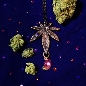 May include: A copper-colored marijuana leaf pendant necklace with a crescent moon charm. The necklace is on a black chain and is surrounded by green marijuana buds and colorful glitter.