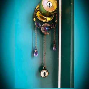 May include: A copper-toned door knocker with a crescent moon design and purple gemstones. The knocker is attached to a chain with a small round pendant featuring a black scorpion.