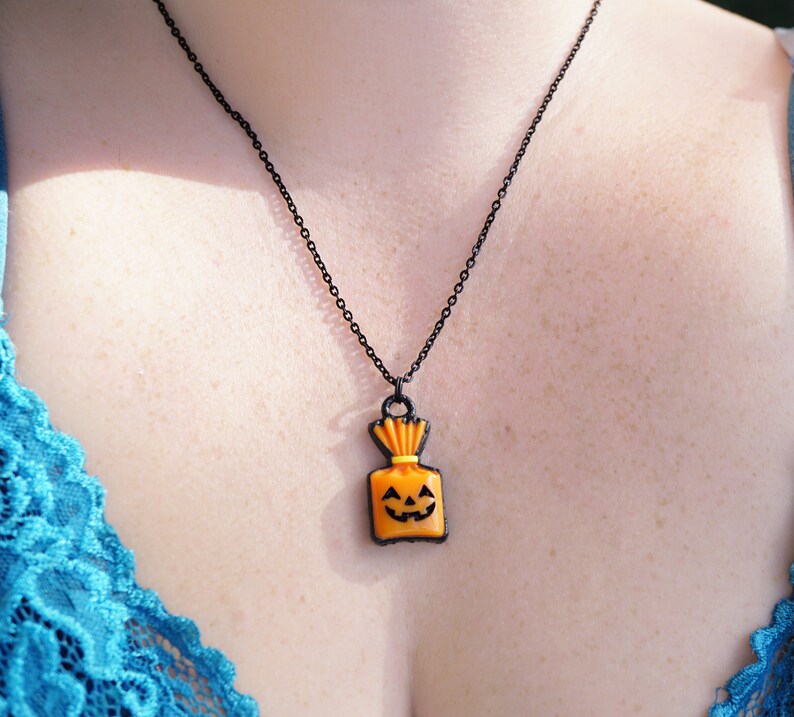 May include: A black chain necklace with an orange pumpkin-shaped pendant. The pendant has a black outline and a smiling jack-o'-lantern face.