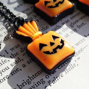 May include: A black necklace with a small orange and black pumpkin-shaped charm. The charm is shaped like a bag with a jack-o-lantern face on the front.