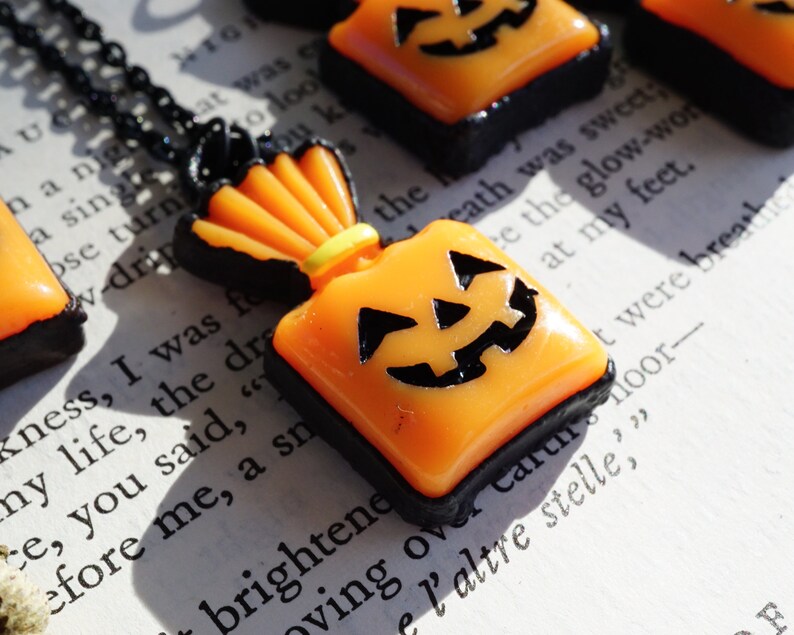May include: A black necklace with a small orange and black pumpkin-shaped charm. The charm is shaped like a bag with a jack-o-lantern face on the front.