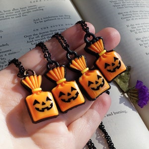 May include: Four black chain necklaces with orange and black pumpkin-shaped candy bag charms. The charms have a yellow string tied around the top and a black outline.