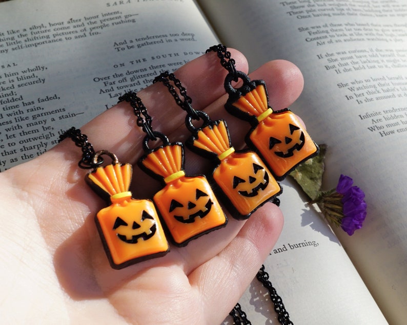 May include: Four black chain necklaces with orange and black pumpkin-shaped candy bag charms. The charms have a yellow string tied around the top and a black outline.