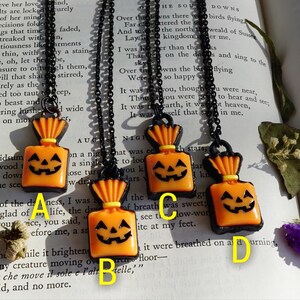 May include: Four orange and black Halloween-themed necklaces with pumpkin-shaped charms. Each charm is a small, orange, fabric bag with a jack-o'-lantern face. The necklaces are on a black book with white text.