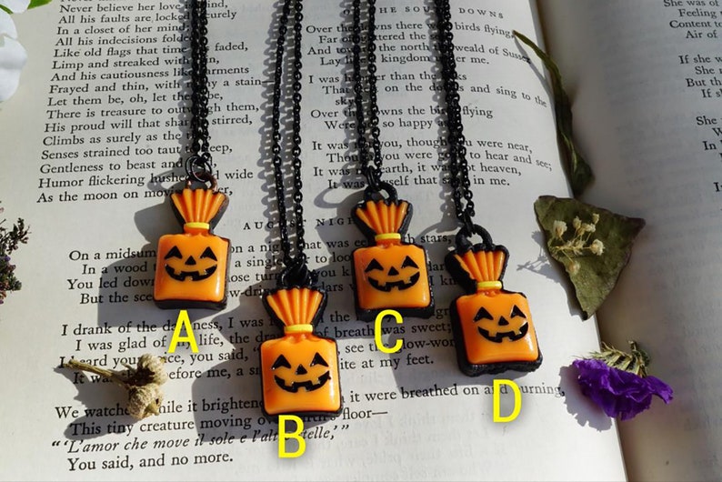 May include: Four orange and black Halloween-themed necklaces with pumpkin-shaped charms. Each charm is a small, orange, fabric bag with a jack-o'-lantern face. The necklaces are on a black book with white text.