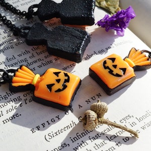 May include: Two Halloween-themed necklaces with black chains. One necklace features a black, 3D, pumpkin-shaped pendant. The other necklace features a bright orange and black candy bag pendant with a jack-o'-lantern face.