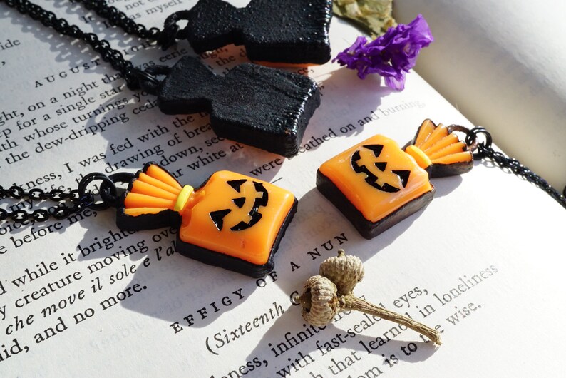 May include: Two Halloween-themed necklaces with black chains. One necklace features a black, 3D, pumpkin-shaped pendant. The other necklace features a bright orange and black candy bag pendant with a jack-o'-lantern face.