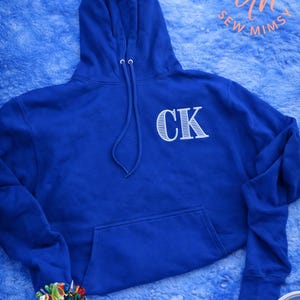 Op de afbeelding: Een koningsblauwe hoodie met een voorzak en een capuchon met witte trekkoorden. De letters "CK" zijn wit op de borst geborduurd. De hoodie wordt gepresenteerd op een blauw, getextureerd oppervlak. De afbeelding bevat ook de tekst "Sew Mimsy".