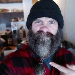 May include: A copper-colored metal beard ring with a twisted wire design, secured around a section of facial hair. The accessory features a metallic finish and a circular shape, positioned centrally on the beard of the individual wearing a black beanie.