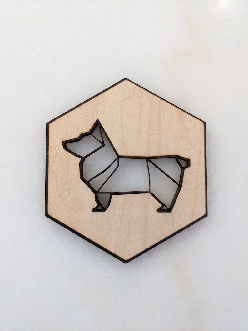 May include: A wooden hexagon-shaped coaster with a laser-cut origami-style corgi dog design. The dog is depicted in shades of gray, set against a light wood background. The coaster has a dark brown border.