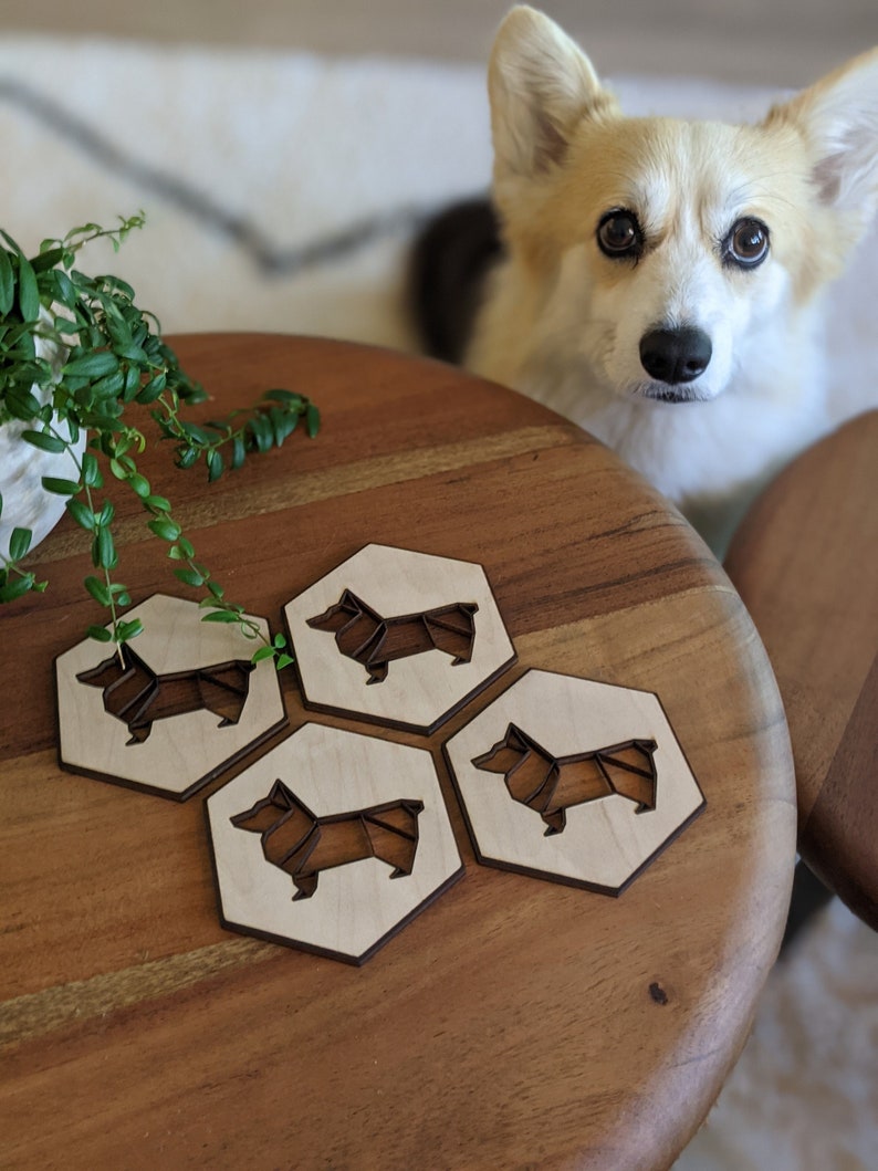 May include: Four hexagonal wooden coasters with a laser-cut corgi dog design. The coasters are light-colored with a darker brown outline and dog silhouette. The coasters are on a wooden table.