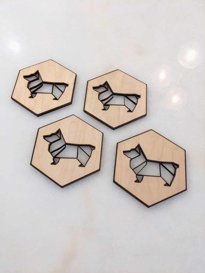 May include: Four wooden coasters with a geometric corgi design. Each coaster is hexagonal with a light wood finish and a dark outline of the corgi. The corgi design is cut out of the wood.